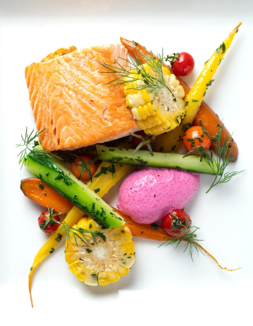 A plate of salmon with colorful vegetables, including corn, carrots, and cucumbers, garnished with herbs and pink sauce.